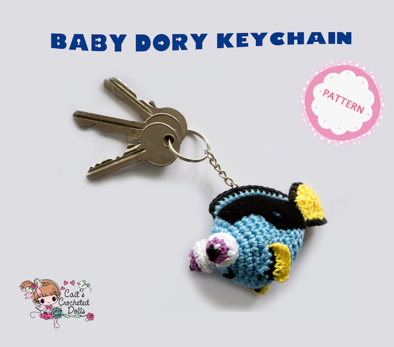 Op de afbeelding: Een gehaakte Baby Dory sleutelhanger in blauw, zwart en geel, bevestigd aan een sleutelring met drie sleutels. De sleutelhanger heeft grote, gedetailleerde ogen en een vinnenontwerp. De tekst bovenaan luidt "BABY DORY KEYCHAIN".