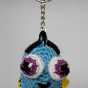 Op de afbeelding: Een gehaakte sleutelhanger met een blauwe vis met grote ogen. De vis heeft witte en paarse ogen, zwarte accenten en gele vinnen. Hij hangt aan een zilveren sleutelring en ketting. Het is een handgemaakt item.