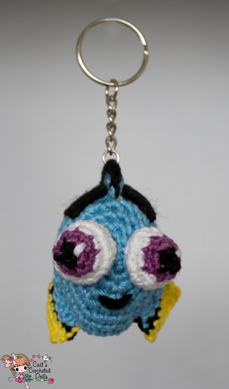 Op de afbeelding: Een gehaakte sleutelhanger met een blauwe vis met grote ogen. De vis heeft witte en paarse ogen, zwarte accenten en gele vinnen. Hij hangt aan een zilveren sleutelring en ketting. Het is een handgemaakt item.