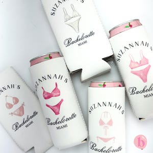 May include: Five white can coolers with pink tops. Each cooler features a pink and white bikini illustration and the text "Suzannah's Bachelorette Miami".