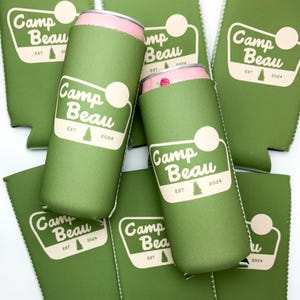 K&ouml;nnte beinhalten: Sechs gr&uuml;ne Neopren-Getr&auml;nkedosenk&uuml;hler mit dem Aufdruck "Camp Beau" und "EST 2024". Die K&uuml;hler sind &uuml;bereinander gestapelt.