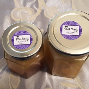 May include: Two glass jars of lavender honey with silver lids. Each jar has a purple label with the text "Chat Noir Lavender Honey". The jars are on a patterned, light-colored surface.