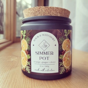 Könnte beinhalten: Ein schwarzes Glas-Kerzengefäß mit einem Korkdeckel. Das Etikett lautet "WICK & WITCHERY" und "SIMMER POT orange-ginger-clove 12 oz. | 340g made with intention". Das Etikett zeigt eine botanische Illustration von Orangen, Nelken und immergrünen Zweigen.