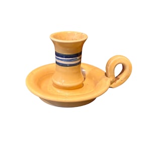 Puede incluir: Un candelero de cerámica con un esmalte amarillo-naranja. El soporte tiene una base circular con un asa y una copa central. La copa está decorada con rayas azules y blancas. El candelero es un objeto decorativo.