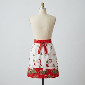 May include: A white apron featuring a repeating pattern of Santa Claus figures emerging from red brick chimneys. The apron includes a bright red fabric waistband that ties in the front with a bow and a red hem decorated with green holly leaves.