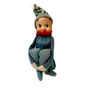 Puede incluir: Una muñeca elfo de estilo vintage con cara de plástico pálido, ojos azules y cuerpo de fieltro azul suave. El elfo lleva un gorro puntiagudo a rayas verdes y blancas con borde gris, un cuello de fieltro rojo y manoplas de fieltro blanco.