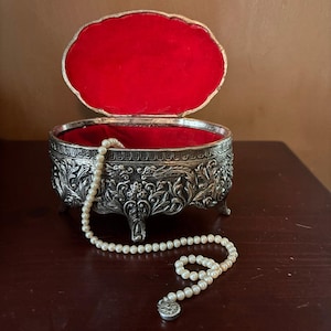 May include: An oval silver-toned metal jewelry box with an intricate embossed floral and scroll pattern, standing on four small feet. The interior is lined with bright red velvet. A single strand of small white pearls rests inside and spills out.