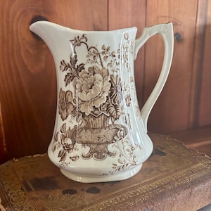 May include: Cream-colored ceramic pitcher with a brown floral design. The pitcher features a large handle and a spout for pouring. The floral pattern includes a basket of flowers and other botanical elements. The pitcher is likely vintage or antique.