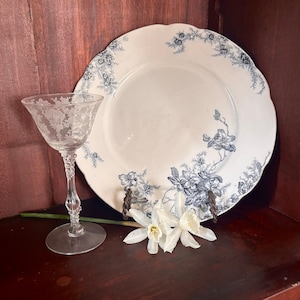 Puede incluir: Un vaso transparente grabado con un diseño floral detallado y un tallo decorativo. Un plato blanco grande con un patrón floral azul y bordes festoneados. Dos flores blancas se colocan frente al plato.