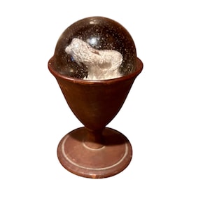 May include: A vintage snow globe featuring a white sculpture inside a clear glass dome. The dome sits atop a brown, goblet-shaped base with a circular foot. The globe has a classic, antique aesthetic.
