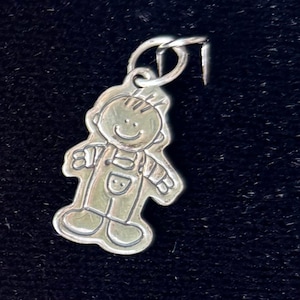 May include: A silver charm depicting a smiling child in overalls. The charm has a loop for attaching to a necklace or bracelet. The charm is set against a dark background.