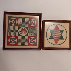 May include: Two framed needlepoint artworks. One features a geometric pattern with red, green, and white accents, and the word "HOME". The other displays a star design in a circle, with red, blue, and tan colors. Both are in brown wooden frames.