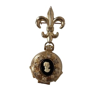 May include: Gold-toned brooch featuring a fleur-de-lis design and a round locket. The locket has a black cameo with a white profile and is surrounded by clear crystals. A vintage-inspired accessory.