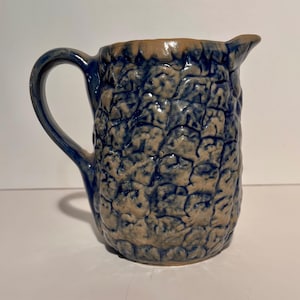 May include: A blue and tan ceramic pitcher with a textured surface. The pitcher has a handle and a spout for pouring. The ceramic pitcher is a decorative item.