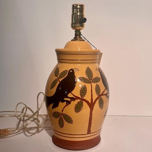 Może przedstawiać: Ceramiczna podstawa lampy stołowej z jasnobrązowym szkliwem typu craquelé. Korpus ozdobiony jest ręcznie malowanymi ciemnobrązowymi ptakami siedzącymi na brązowych gałęziach z zielonymi liśćmi. Posiada mosiężną oprawkę metalową oraz przezroczysty przewód.