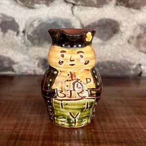 Könnte beinhalten: Handbemalter Keramikkrug in Form einer Person, mit braunem Hut, Jacke und Schürze. Das Gesicht ist beige mit schwarzen Details. Die Schürze trägt die Buchstaben "B.I.R.D." und ein Uhrendesign. Der Krug steht auf einer Holzoberfläche.