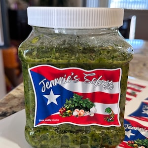 May include: A glass jar filled with green sofrito, labeled "Jeannie's Sofrito" with a Puerto Rican flag design. The label also includes the text "@ms.crafty_mama".