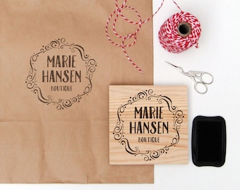 Könnte beinhalten: Eine braune Papiertüte mit einem Holzstempel, auf dem "Marie Hansen Boutique" in einem dekorativen Rahmen steht. Der Stempel befindet sich auf einem Holzblock und daneben liegt ein schwarzes Stempelkissen. Außerdem gibt es eine Spule mit rotem und weißem Garn und eine silberne Schere.