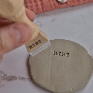 May include: A wooden stamp marked "MINT" is pressed onto a small, round piece of clay. The clay is light brown, and the stamp is being held. Pottery tools are visible in the background, on a terracotta-colored cloth.