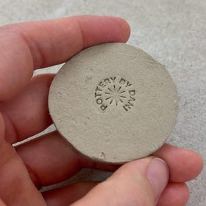 May include: A small, circular, pale grey ceramic piece with the words "POTTERY BY DAN" stamped on it. The text surrounds a starburst design. The piece is held in a person's hand.