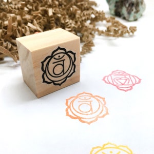 Op de afbeelding: Een houten blokstempel met een zwart-wit afbeelding van het sacrale chakrasymbool, een gestileerde lotusbloem met het Sanskriet-symbool "Svadhisthana" erin. De stempel staat op een wit oppervlak met drie afdrukken van het chakrasymbool in oranje, roze en geel.