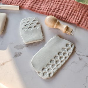 Könnte beinhalten: Eine Sammlung von Töpferwerkzeugen und Tonstücken. Zwei graue Tonfliesen weisen ein sich wiederholendes Jakobsmuschelmuster auf. Ein Holzwerkzeug mit rundem Griff und ein kleines, weißes rechteckiges Tonstück sind ebenfalls zu sehen. Der Hintergrund umfasst ein rosa Tuch und eine grüne Schüssel.