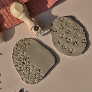 Può includere: Strumenti per la ceramica, tra cui un timbro con manico in legno e due timbri in argilla grigia con motivi floreali. Gli strumenti sono disposti su una superficie rosa chiaro, con un panno rosa sullo sfondo.