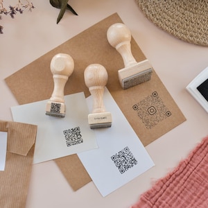 Può includere: Timbri in legno con codici QR sono disposti su una superficie beige chiaro. I timbri hanno un manico arrotondato e una base rettangolare. Sono visibili anche diversi fogli di carta e un sacchetto di carta marrone con la scritta "Crochet is my CARDIO".