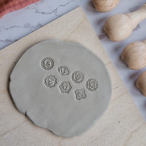 Pode incluir: Uma placa de argila cinza claro com sete símbolos de chakra estampados, repousando sobre uma tábua de madeira. Várias ferramentas com cabo de madeira estão dispostas nas proximidades. Uma tigela verde e um pano rosa estão no fundo.