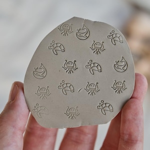 May include: A gray, irregularly shaped clay stamp with an assortment of sea creature designs. The stamp features etched images of turtles, jellyfish, and whales. The stamp is held in a person's hand.