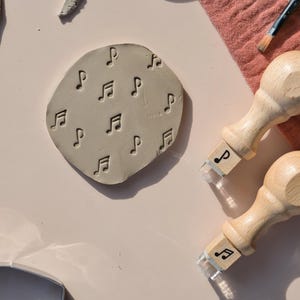 May include: A circular piece of light gray clay featuring an embossed pattern of musical notes. Two wooden stamps with matching musical note designs on their bases are positioned nearby, alongside a paintbrush and a metal cutter tool.