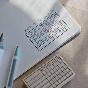 Op de afbeelding: Open notitieboek met een gewoonte-tracker voor Week 4, 26 januari, met vinkjes voor lezen, meditatie, tekenen en water drinken. Een grijze en witte marker en een turquoise pen staan links. Een houten stempel met een lege gewoonte-tracker staat onderaan.
