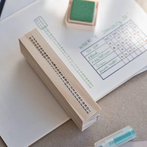 May include: A rectangular wooden date stamp featuring a long vertical sequence of numbers from 0 to 31 printed in black ink on its side. The stamp is positioned on a white paper sheet containing a handwritten weekly planner grid and a green ink pad.