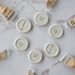 May include: A collection of small, round, off-white ceramic stamps with various phrases in black, including "BE JOYFUL," "LOVE YOU," and "LET IT BE." Each stamp has a wooden handle and a clear plastic base. The stamps are arranged on a marble surface.