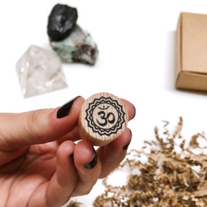 Op de afbeelding: Een houten stempel met een zwart Om-symbool in een decoratieve rand wordt in een hand gehouden. De stempel is rond en gemaakt van lichtgekleurd hout. Andere items in de afbeelding zijn kristallen en een kleine kartonnen doos.