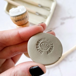 以下が含まれることがあります： グレーの粘土製のスタンプのクローズアップ。スタンプには「Bitterswit Pottery」という文字と貝殻のデザインが刻まれています。スタンプは手で持たれています。