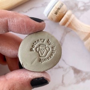 May include: A close-up of a gray clay stamp with the text "Pottery by bitsywife" and a small illustration of a vase with a heart inside.