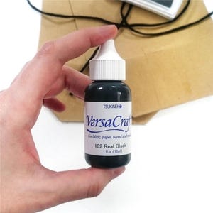 Könnte beinhalten: Eine kleine Flasche schwarzer Tinte mit der Aufschrift "VersaCraft" für Stoff, Papier, Holz und mehr. Die Flasche ist mit "182 Real Black" beschriftet und enthält 1 fl. oz. (30 ml) Tinte.