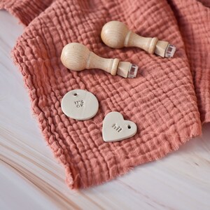 May include: Two wooden stamps with handles and a light brown fabric background. The stamps are used to create personalized tags. The tags are made of white clay and have the words "It's OK" and "Hi!" written on them.