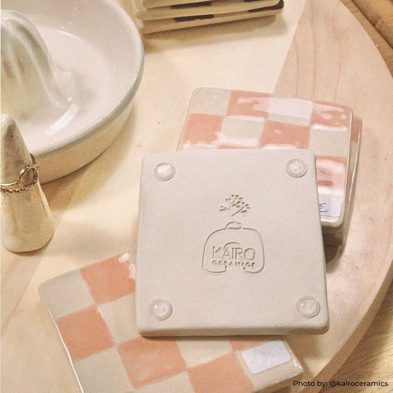 Może przedstawiać: Cztery ceramiczne podkładki pod kubki w r&oacute;żowo-białe kratki. Jedna podkładka ma białe logo z tekstem "KAIR0 ceramics" i małą ilustrację rośliny.