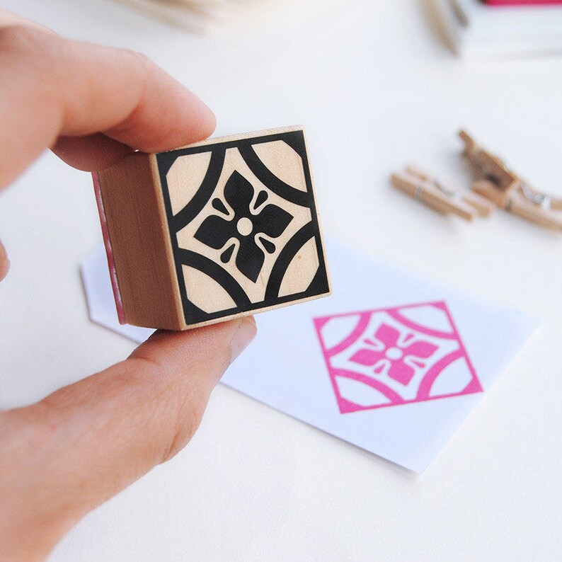 May include: A wooden stamp with a black and white geometric design of a flower. The stamp is being held in a hand and a pink impression of the design is visible on a white surface.
