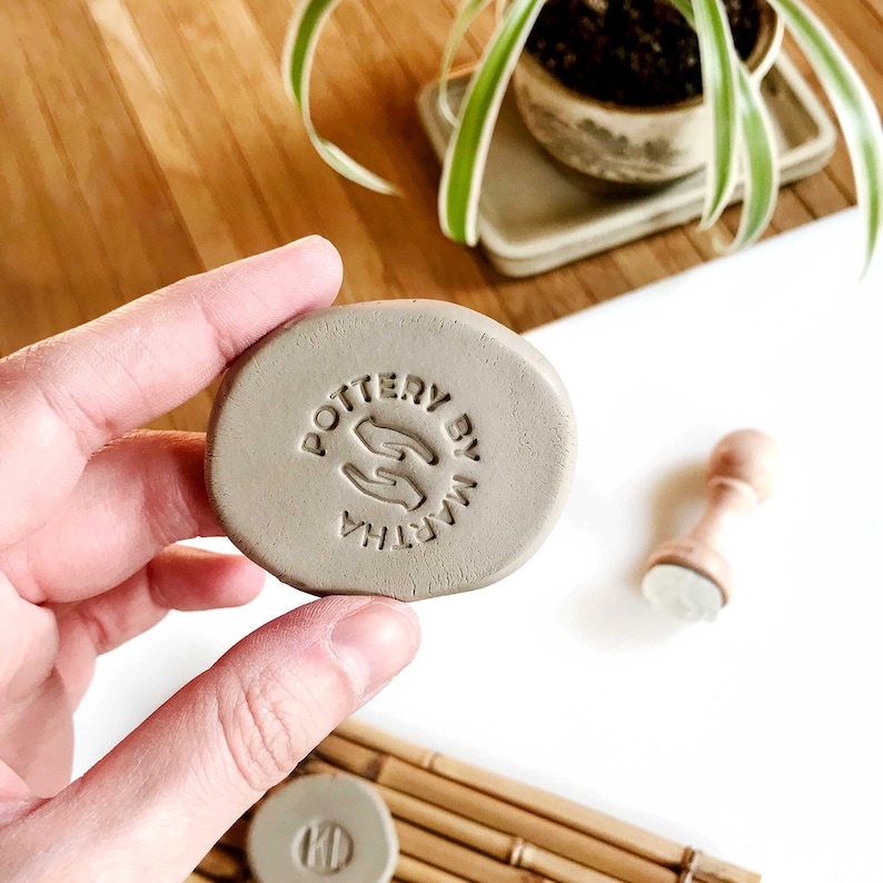 May include: A close-up of a gray clay stamp with the text "POTTERY BY MARTHA" and a graphic of two hands holding a heart. The stamp is being held by a hand.