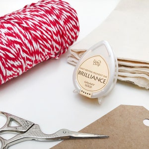 May include: A white ink pad with the text "Dew Drop Brilliance Fast-Drying Pigment Ink Galaxy Gold" on the front. The ink pad is sitting on a white surface next to a roll of red and white twine, a piece of white fabric, and a brown tag.