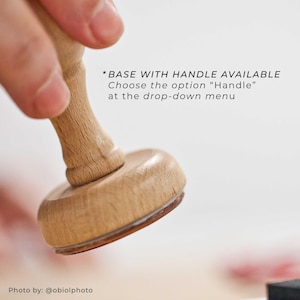 Op de afbeelding: Een houten stempel met een afgeronde basis en handvat. De basis heeft een bruine rand. Tekst op de afbeelding luidt: "*BASE WITH HANDLE AVAILABLE Choose the option "Handle" at the drop-down menu."