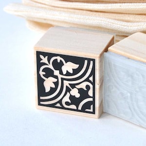 Puede incluir: Un sello de madera con un dise&ntilde;o en blanco y negro de un patr&oacute;n repetitivo de hojas y espirales. El sello est&aacute; montado en un bloque de madera.