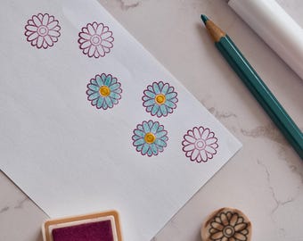 Könnte beinhalten: Ein weißes Blatt Papier mit mehreren gestempelten Gänseblümchen-Umrissen in dunkelvioletter Tinte, wobei einige Blüten hellblau und gelb ausgemalt sind. Ein dunkelgrüner Buntstift, ein weißer Marker, ein violettes Stempelkissen und ein runder Stempel.