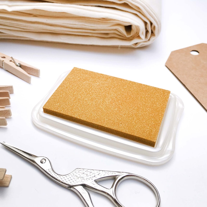 May include: A gold ink pad with a white plastic case. The ink pad is on a white surface with a pair of small scissors and a stack of fabric.