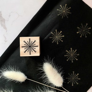 May include: A wooden stamp with a black snowflake design on a black fabric background with gold snowflakes printed on it.
