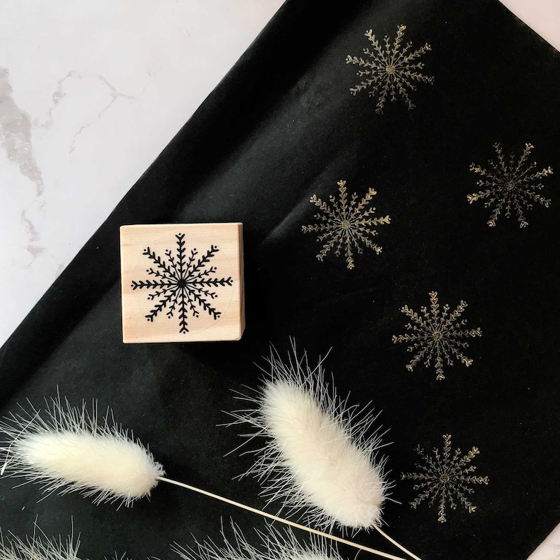 May include: A wooden stamp with a black snowflake design on a black fabric background with gold snowflakes printed on it.
