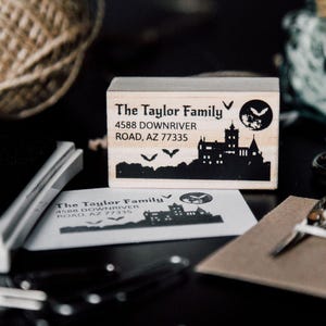 On a moody dark background, our Transylvania-themed return address rubber stamp is printed on a white card, surrounded by a notebook, black scissors, and themed stationery. The stamp features bats and a full moon, perfect for Halloween mail or gothic fantasy fans. This dark stationery setup complements the moody aesthetic of the floral ex libris stamp—an ideal match for book lovers who live for storytelling vibes and spooky mail art.